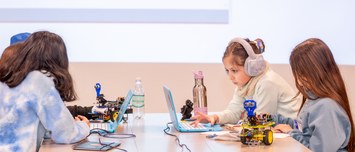 Una niña en una habitación rosada, con audífonos mirando la pantalla de un computador. A su lado hay un robot pequeño.