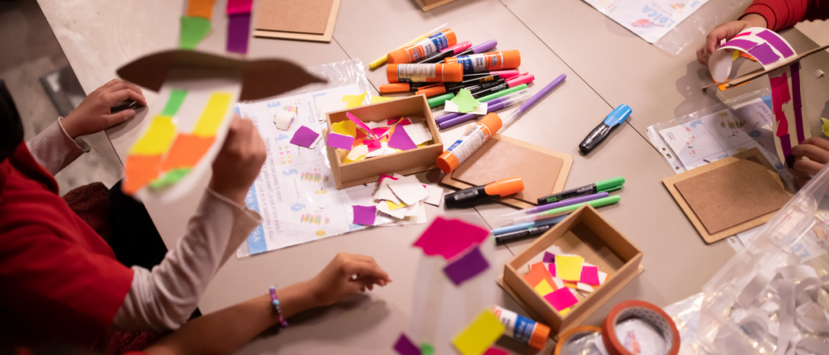 Vemos las manos de un grupo de niños y niñas haciendo manualidades en una mesa grande. En la mesa se ven muchos materiales, texturas y colores.