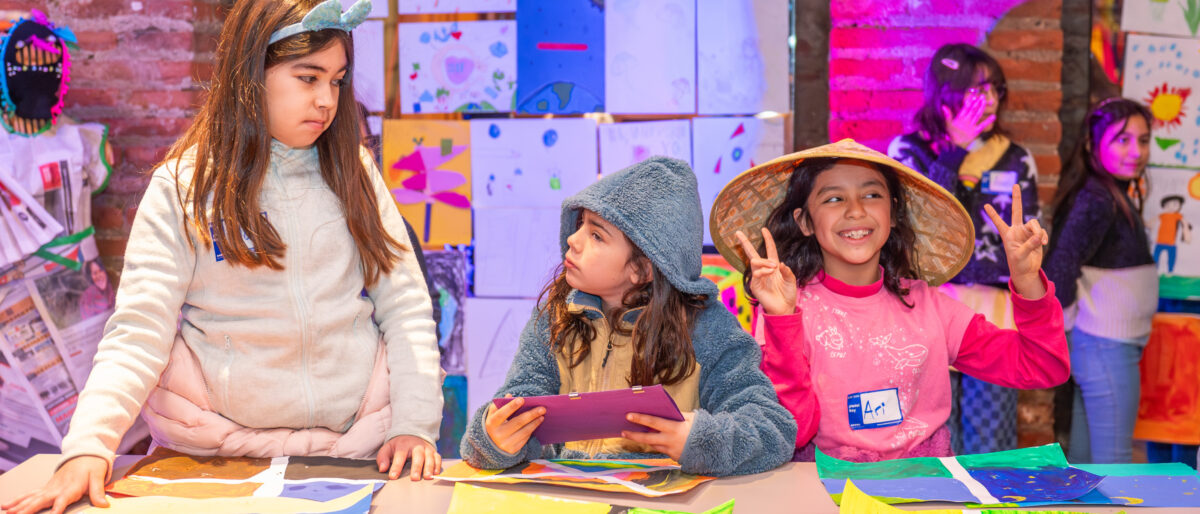 Grupo de niñas que están aprendiendo la importancia de jugar por jugar. Las vemos junto a papeles de colores, probándose sombreros y conversando en un ambiente distendido.